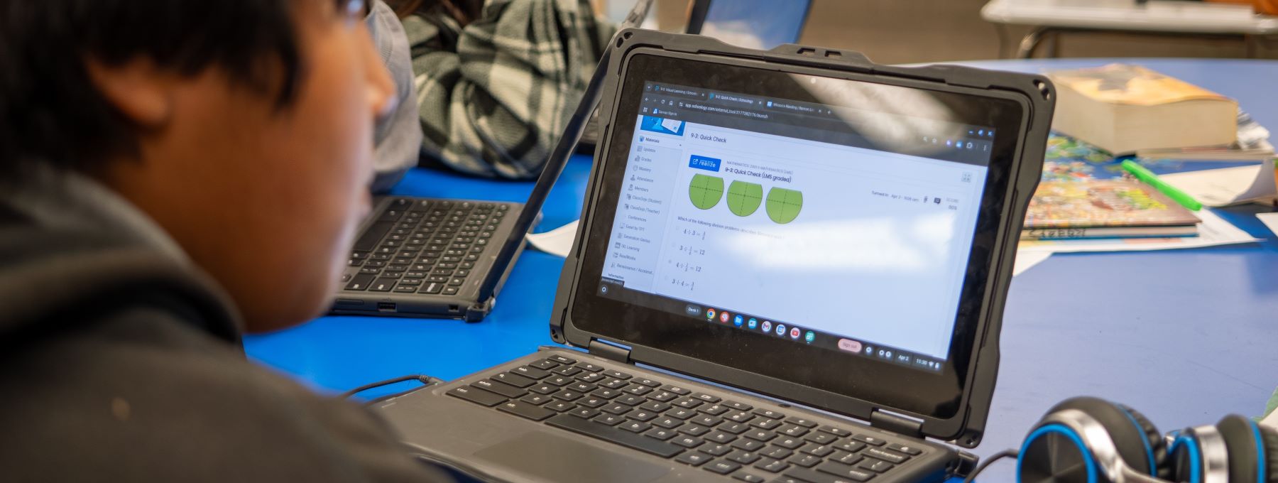 A student focused on a laptop in a bright classroom, surrounded by books and classmates engaged in learning.