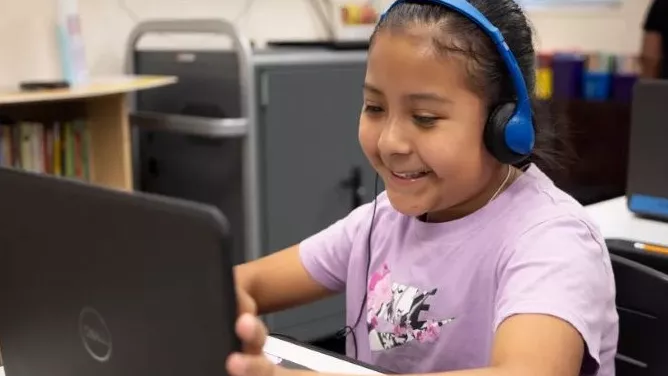 A student wearing headphones smiles while focused on laptop screen, engaged in an online activity.